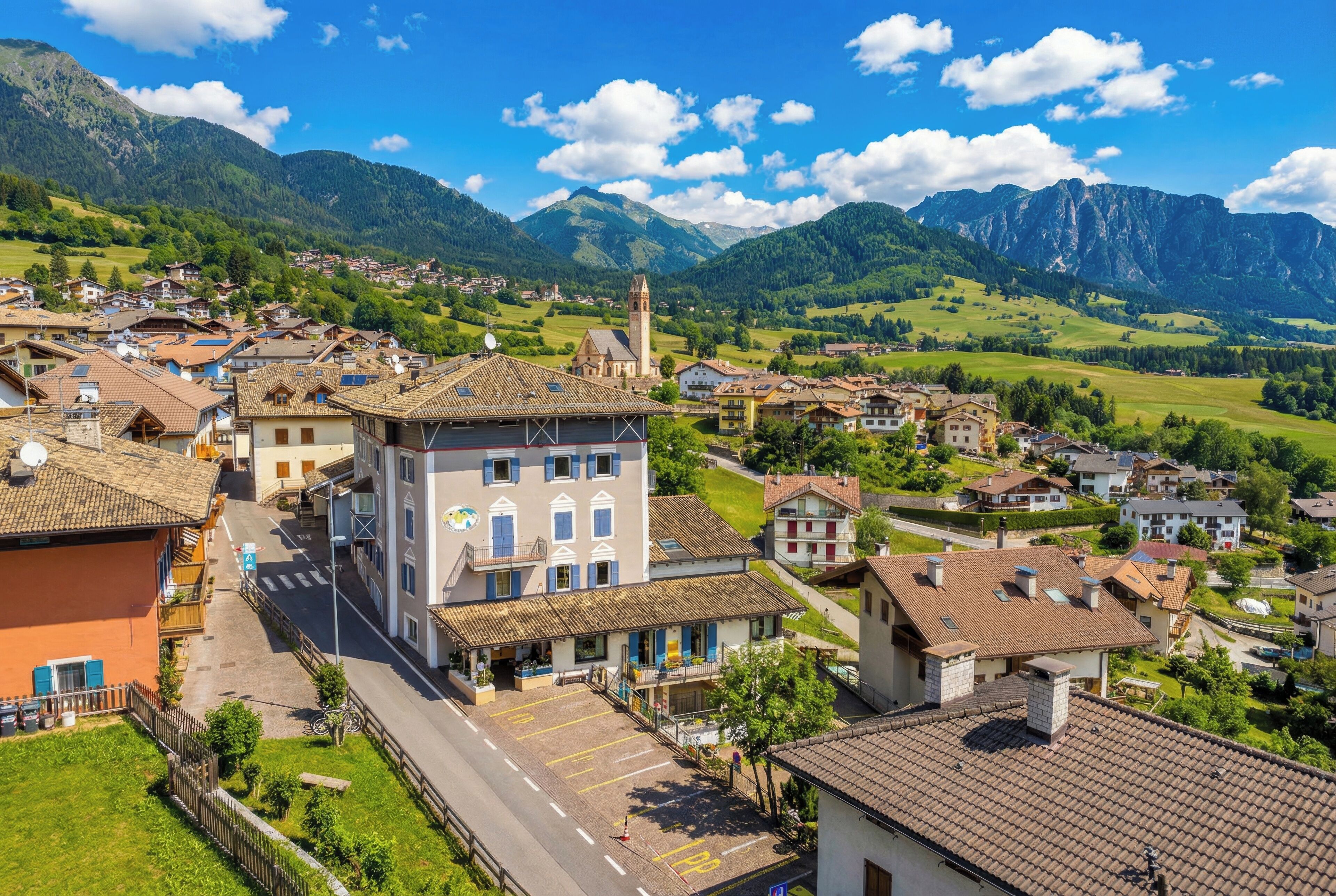 A town with a prominent building featuring a tower, surrounded by residential houses and lush green hills.