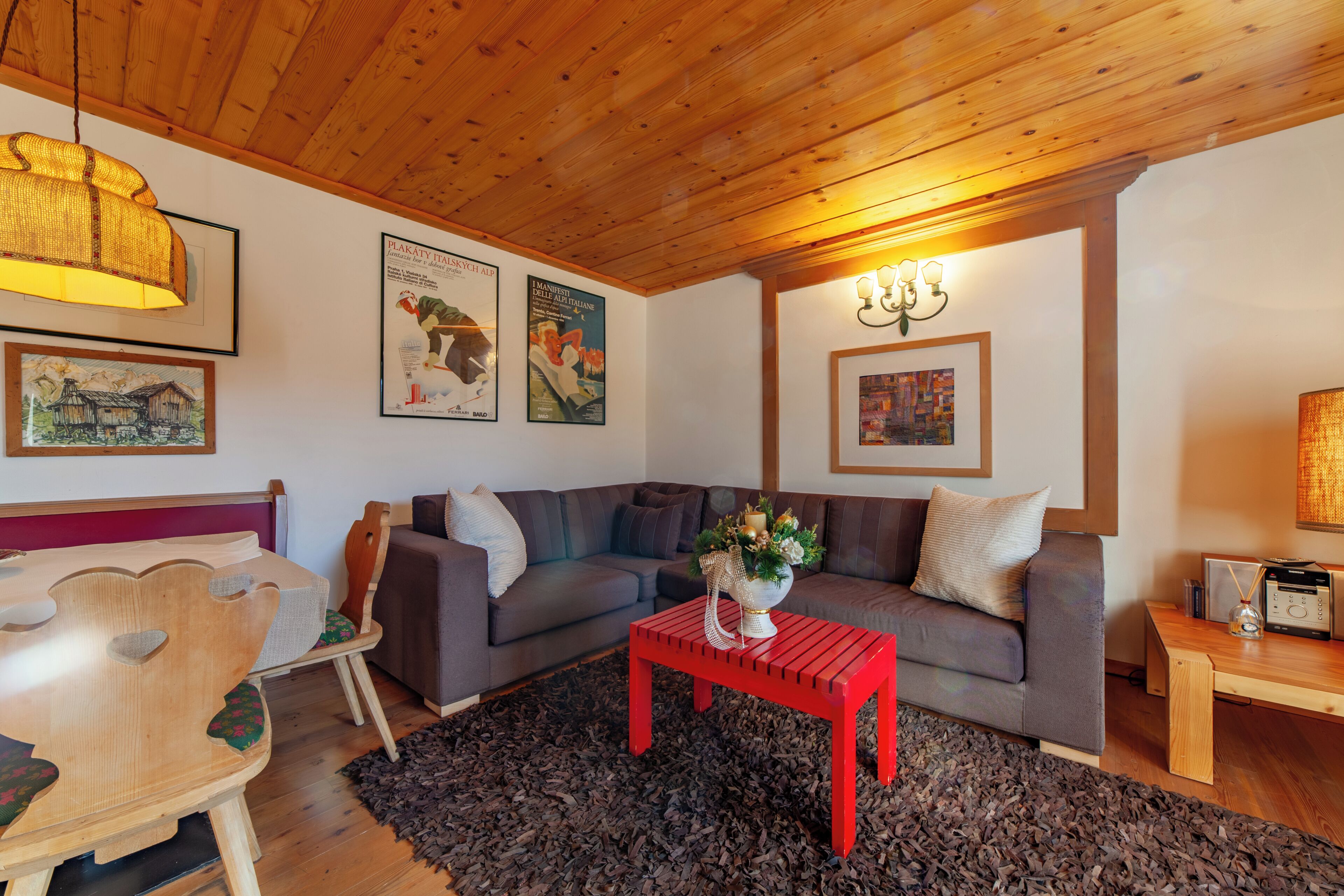 A cozy living room with a wooden ceiling, a grey sofa, a red coffee table, and framed artwork on the walls.