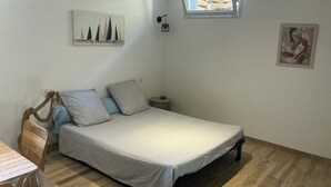 Quarto com cama, mesa de cabeceira pequena, prateleira na parede com um quadro, janela e luminária de teto.