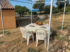 Un espace de restauration en plein air avec une table dressée pour deux personnes, située sous une pergola blanche, et entourée d’herbe sèche et d’un mur de pierre.