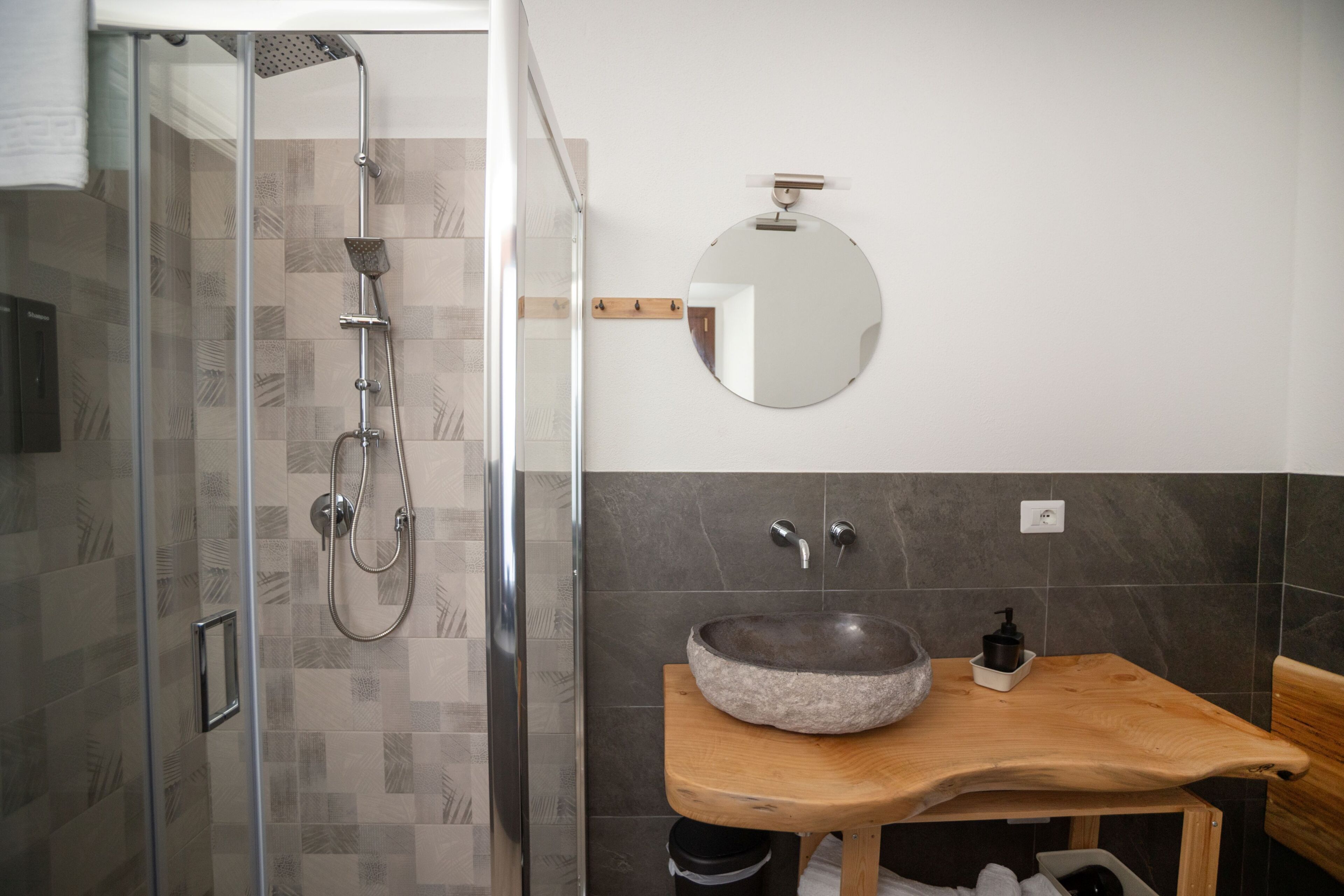 Ein Badezimmer mit Duschkabine aus Glas, einem Holzwaschtisch mit Steinbecken, einem runden Spiegel und einem an der Wand befestigten Handtuchhalter.