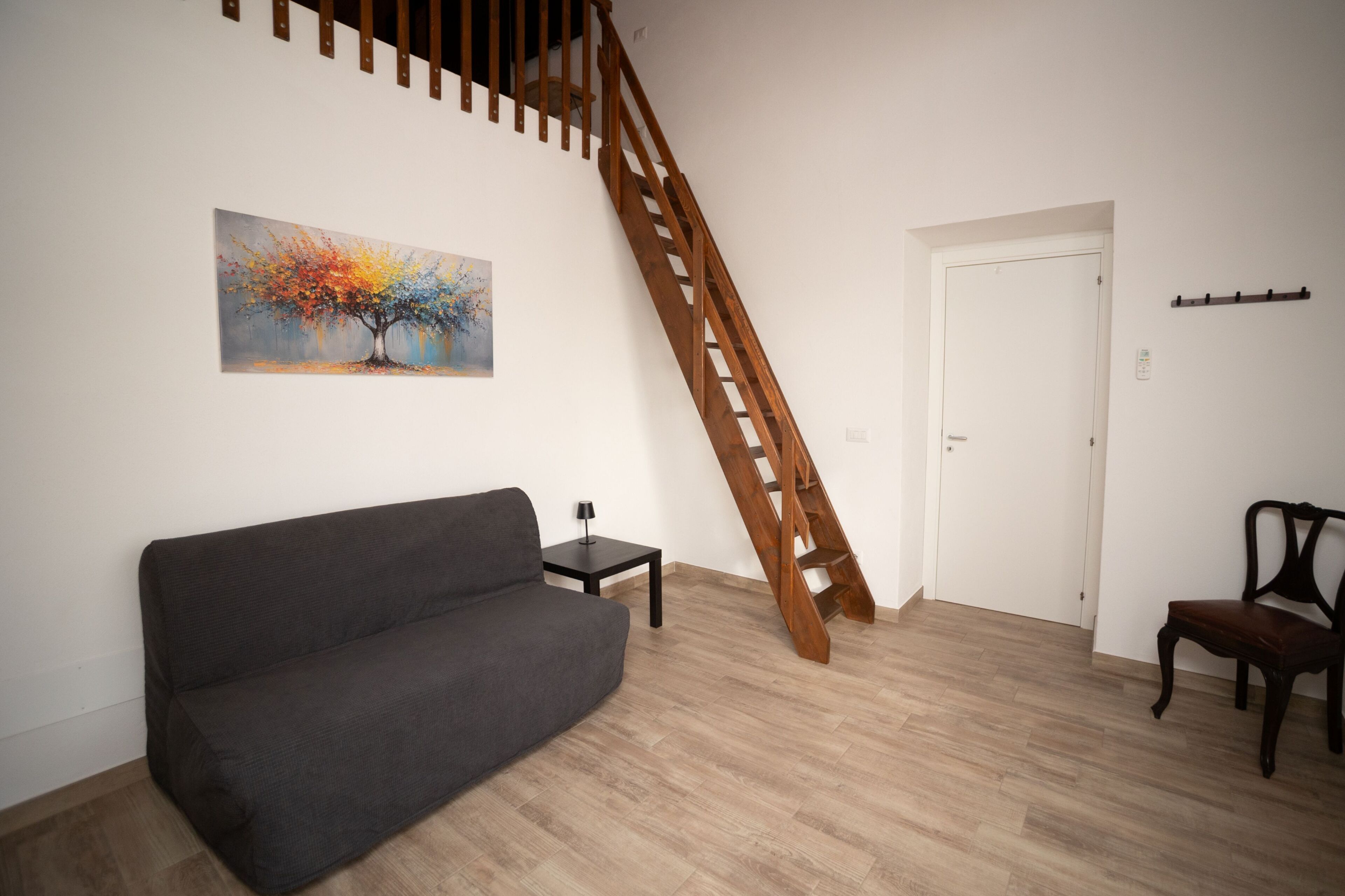 Ein Wohnzimmer mit grauem Sofa, einem kleinen Tisch mit Lampe, einer Holztreppe und einer Tür.