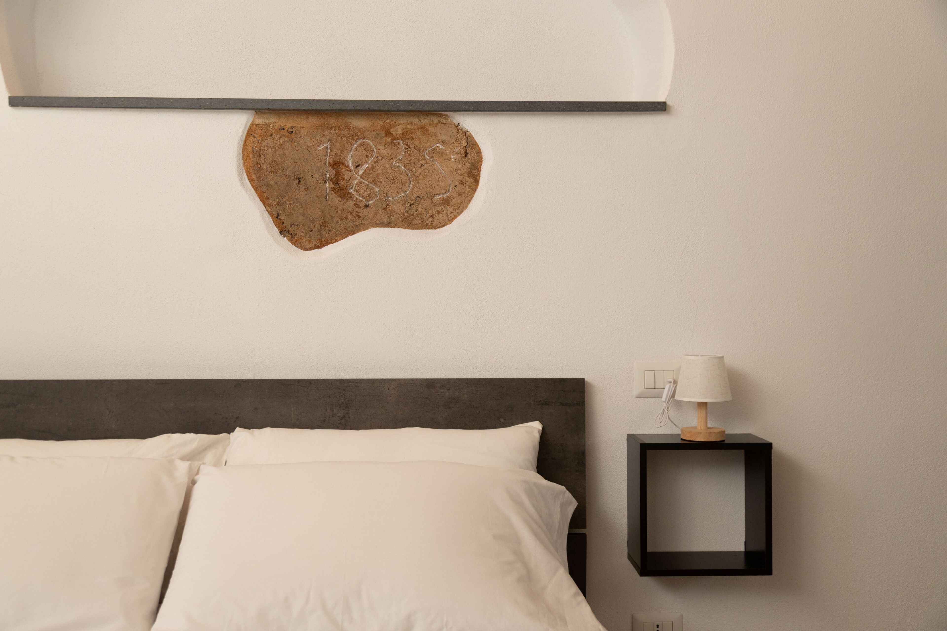 Una cama con ropa de cama blanca, un cabecero oscuro, una lámpara de pared y una mesita de noche con lámpara.