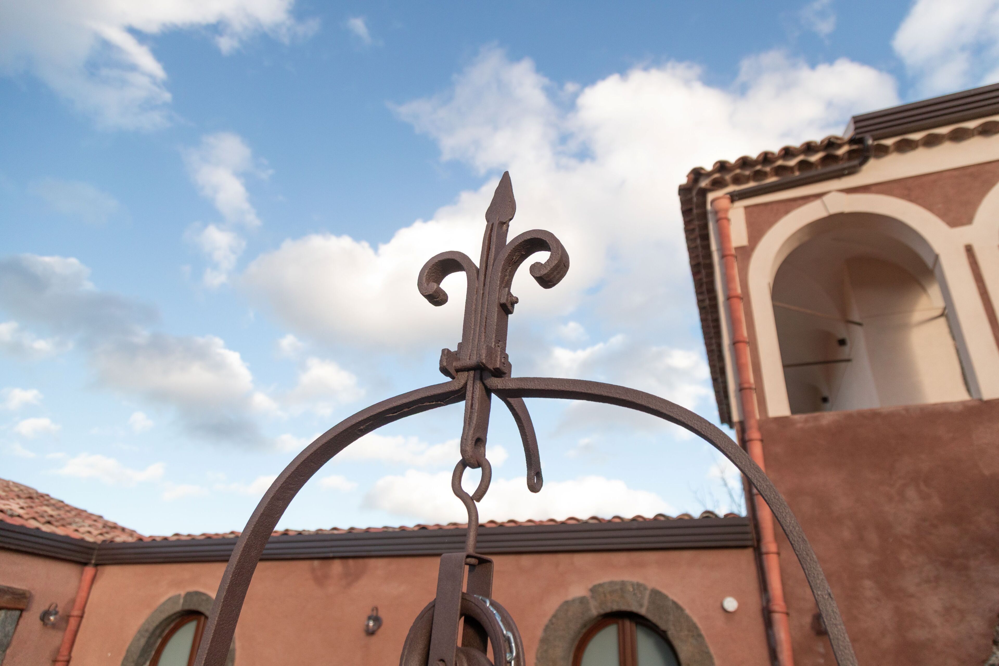 Une porte en fer forgé ornée d’un motif en forme de fleur de lys, qui se dresse devant un bâtiment doté d’arcatures et d’un toit recouvert de tuiles.