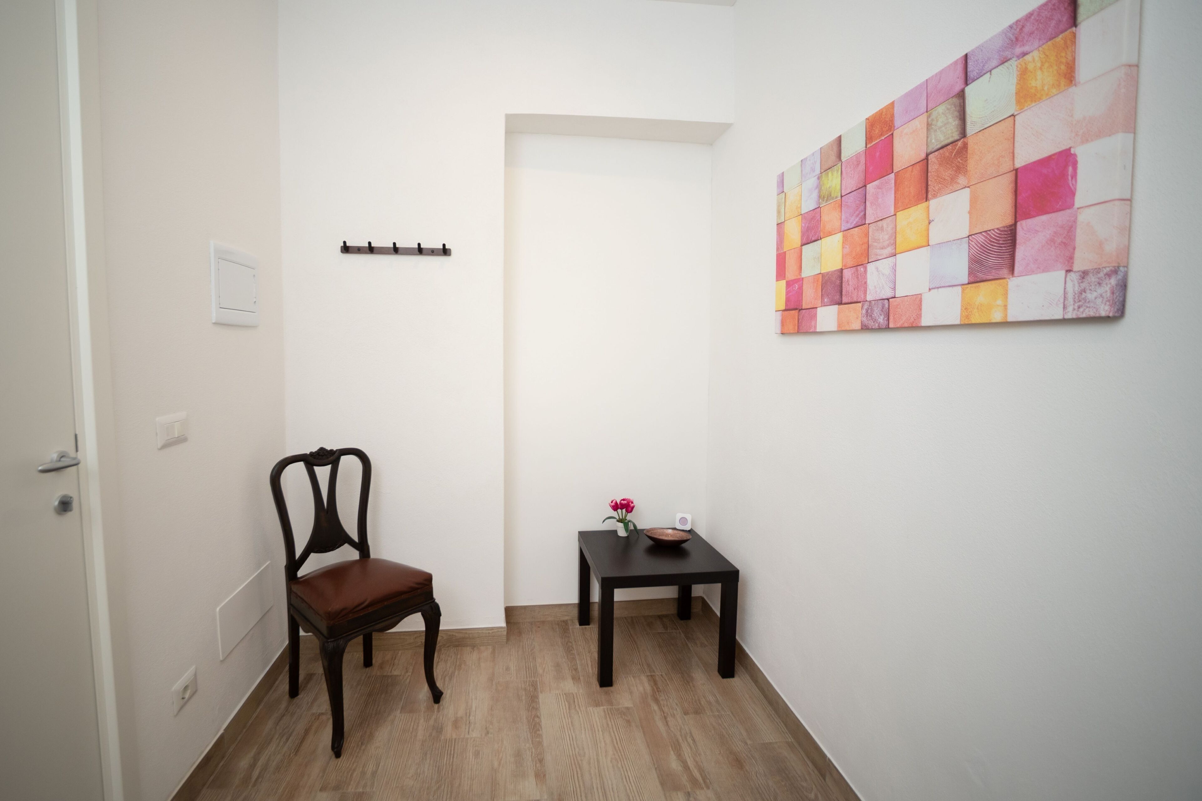 A hallway with a wooden chair, a small table with a vase and a bowl, and a colorful abstract painting on the wall.