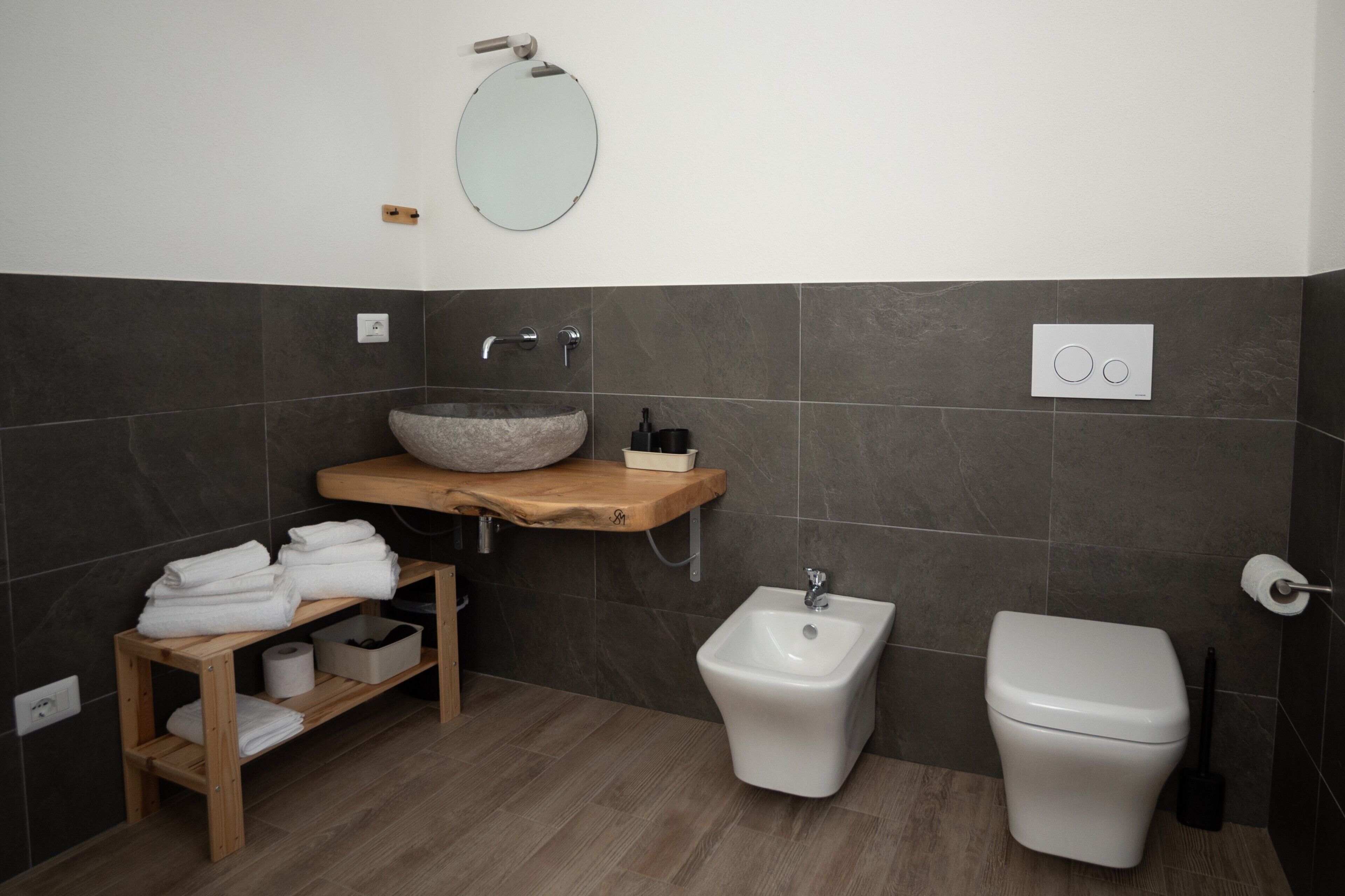 Un baño moderno con tocador de madera, un espejo redondo, un bidé y un inodoro.