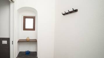 Ein Badezimmer mit weißer Wand, einem Fenster, einem Regal mit einer Pflanze und einem Kleiderhaken.