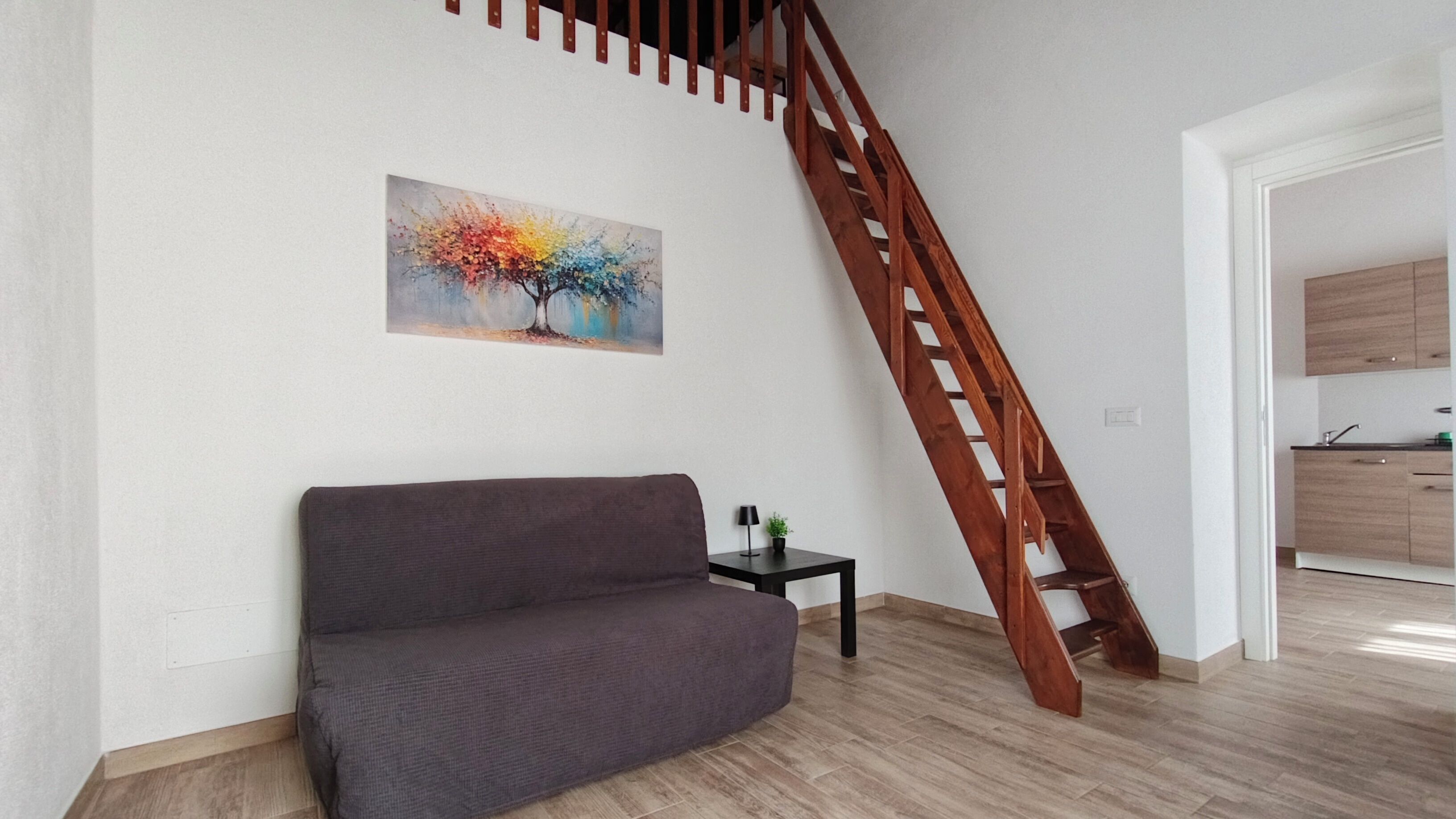 Una sala de estar con un sofá, un cuadro, una mesita con una lámpara y una escalera de madera.