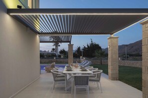 Um pátio externo com mesa posta para refeições, uma pérgola acima e vista para uma piscina e montanhas ao fundo.