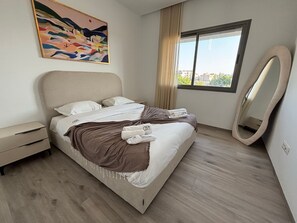 Ein modernes Hotelzimmer mit Bett, Nachttisch, einem großen Spiegel und einem Fenster mit Blick auf Gebäude.