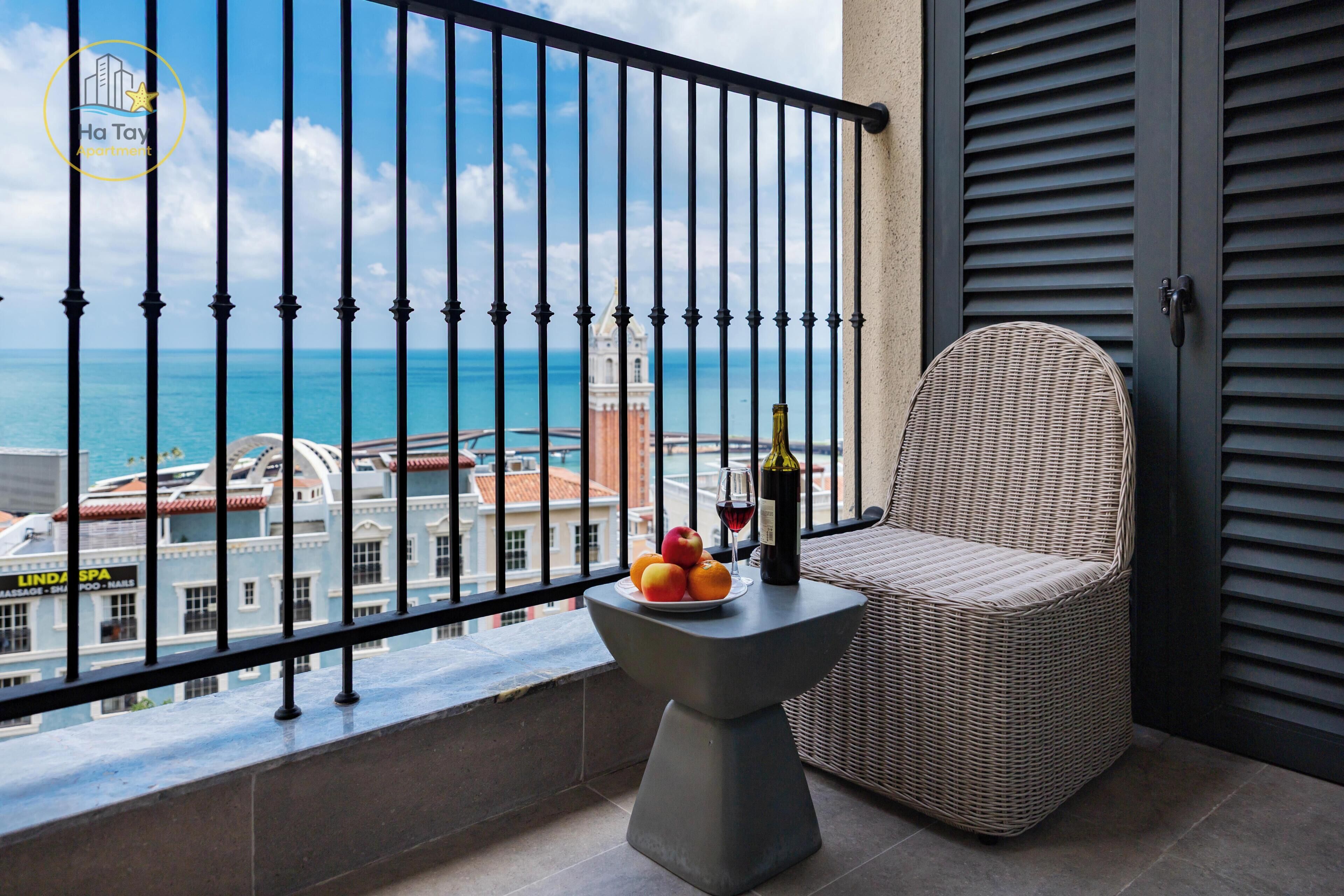 A balcony with a wicker chair, a small table with fruit and a bottle, and a view of the sea and buildings.