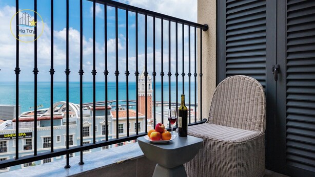 Un balcon avec un fauteuil en osier, une petite table avec des fruits et une bouteille, et une vue sur la mer et les bâtiments.