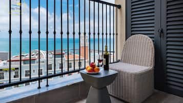A balcony with a wicker chair, a small table with fruit and a bottle, and a view of the sea and buildings.
