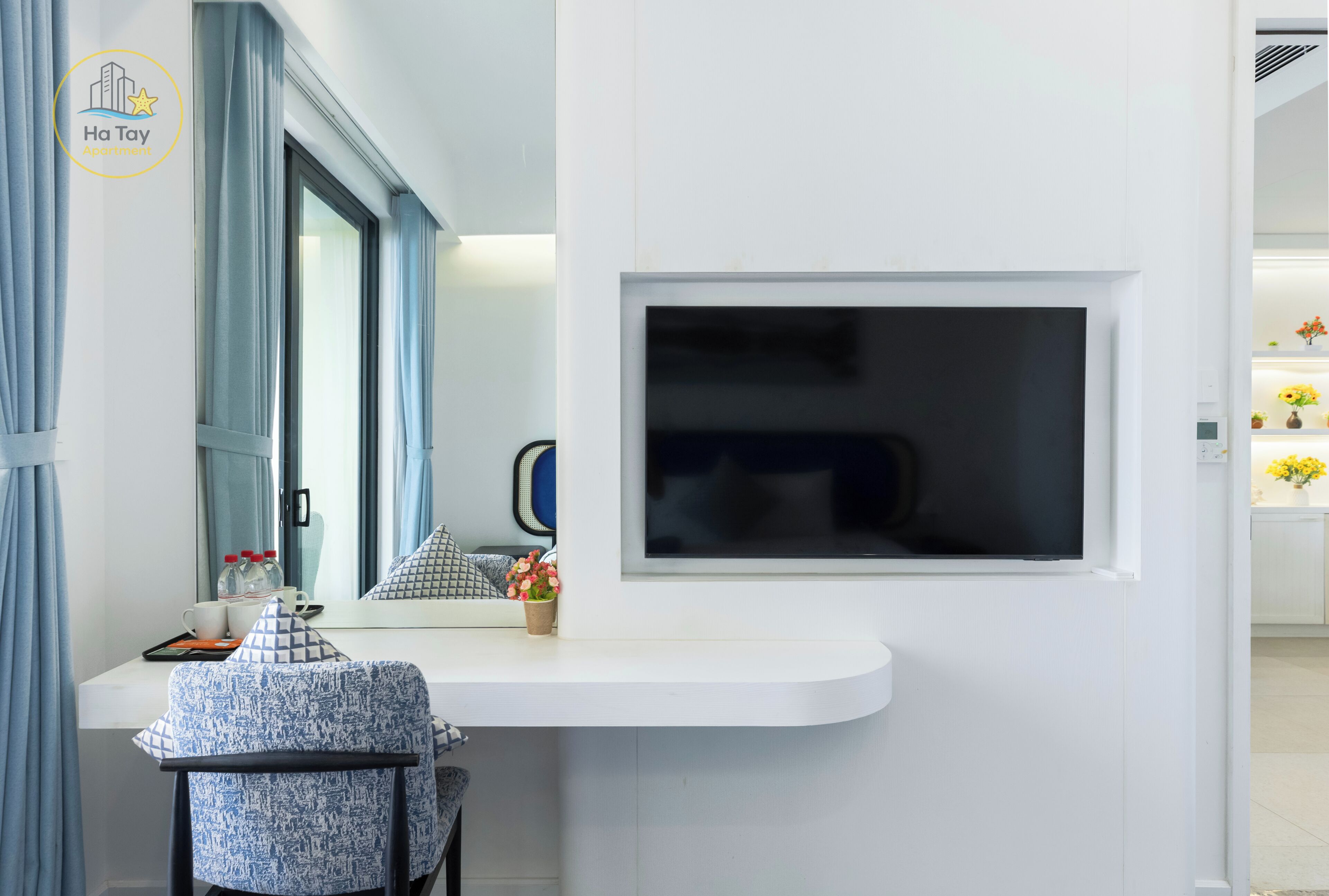 A modern room with a white desk, a flat-screen TV mounted on the wall, a window with curtains, and a small potted plant.