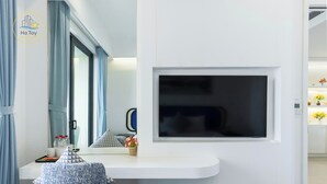 Une chambre moderne avec un bureau blanc, un téléviseur à écran plat fixé au mur, une fenêtre avec des rideaux et une petite plante en pot.