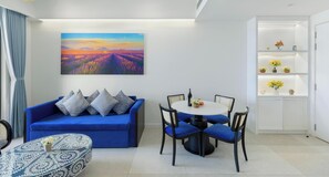 A modern living room with a blue sofa, a round dining table with chairs, a painting of a landscape, and a white shelving unit with decorative items. - One Bedrooms Apartment With Sea View (Phu Quoc)