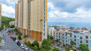 Un immeuble de grande hauteur à la façade jaune, avec vue sur la mer, et une rue bordée de voitures garées et d’arbres.