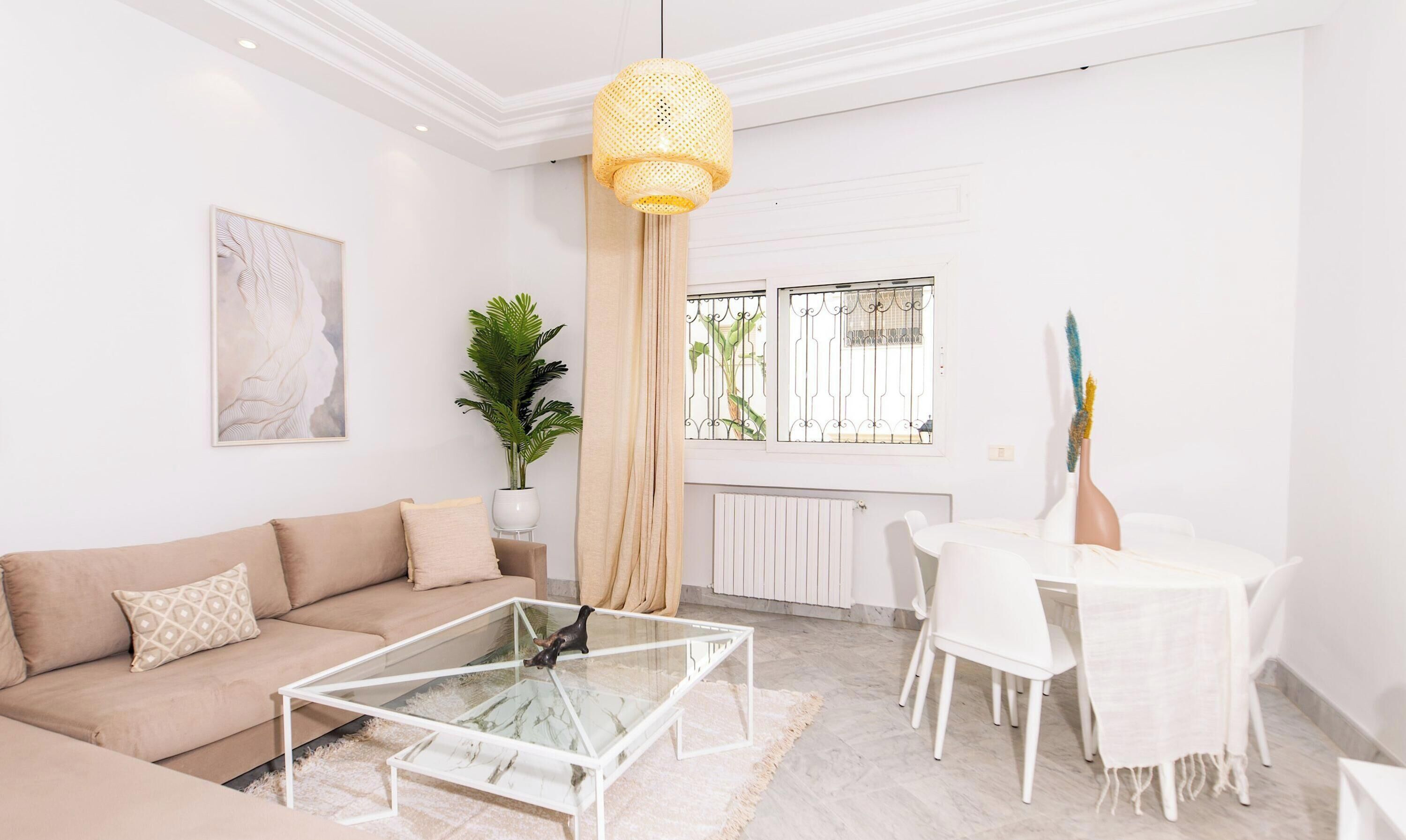 A living room with a sofa, a glass coffee table, a dining table with chairs, a potted plant, and a window with metal bars.