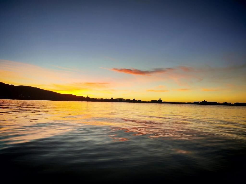 A calm body of water reflecting a sunset with silhouettes of buildings on the horizon.