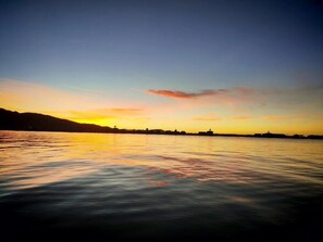 A calm body of water reflecting a sunset with silhouettes of buildings on the horizon.