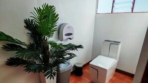 Un baño pequeño con inodoro, un cubo de basura, una planta en maceta y una ventana.