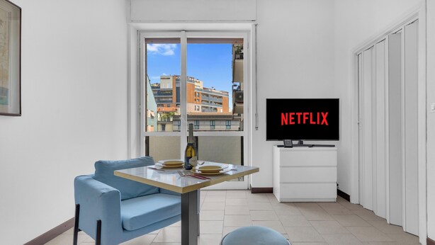 En moderne stue med en blå sofa, et spisebord med tallerkener og bestik, en vinflaske og et fjernsyn, der viser 'NETFLIX' i røde bogstaver.