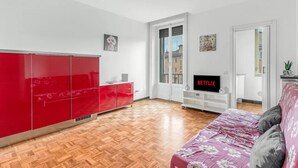 Habitación moderna con piso de madera, un armario rojo, un mueble blanco para televisión y una cama con colcha estampada.