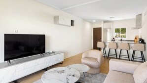 Ein modernes Wohnzimmer mit Fernseher, Sofa und Couchtisch, eine Küche mit Barhocker und ein großes Fenster.