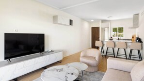 Ein modernes Wohnzimmer mit Fernseher, Sofa und Couchtisch, eine Küche mit Barhocker und ein großes Fenster.