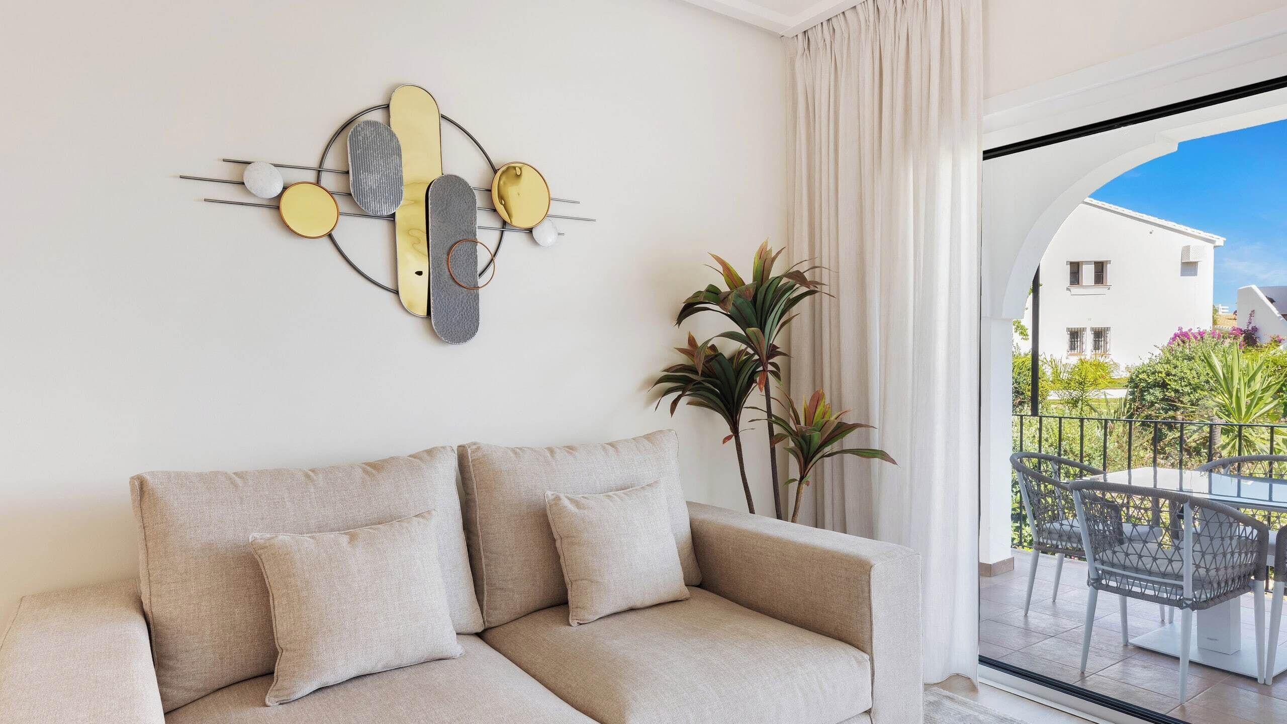 Een moderne woonkamer met een bank, een abstract kunstwerk aan de muur, en uitzicht op een balkon met tuinmeubilair.