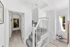 Eine Treppe mit weißen Geländern führt zu einer oberen Etage, eine Tür mit grünem Ausgangsschild und ein Fenster mit Blick auf Grünflächen.