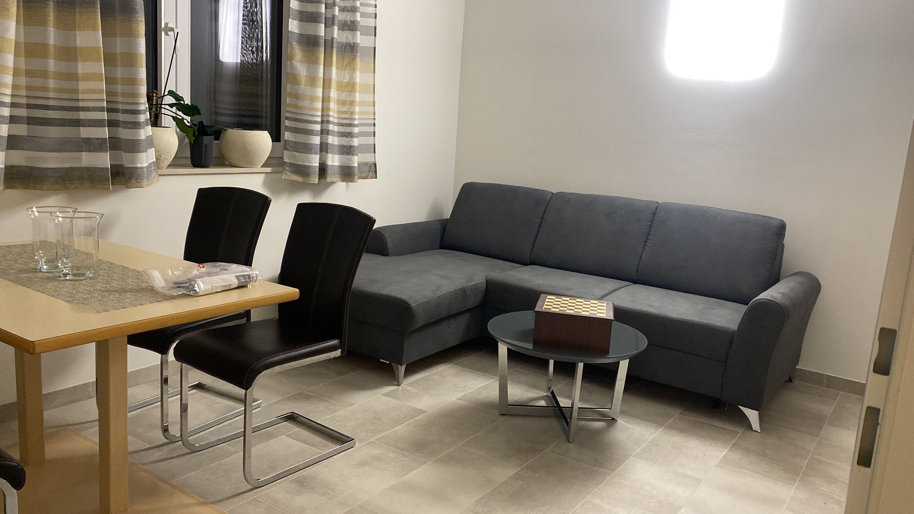 Un salon moderne avec un canapé d’angle gris, une petite table basse ronde, une table à manger avec deux chaises et une fenêtre avec des rideaux.