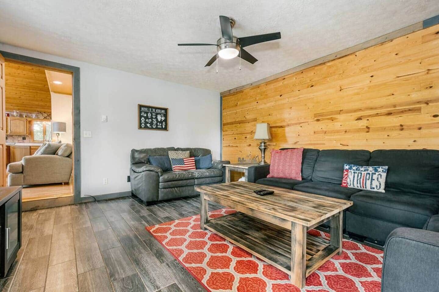 Una sala de estar con una pared de madera, un ventilador de techo, una chimenea y un tapete rojo estampado.