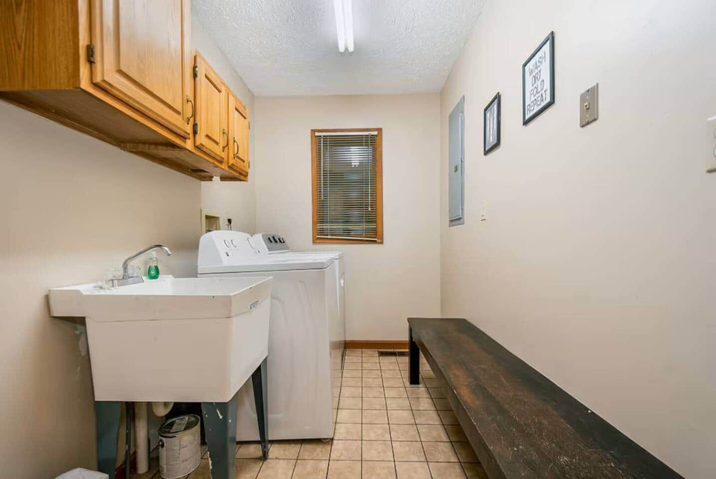 A laundry room with a sink, washing machines, a bench, and a window with blinds.