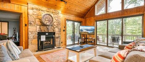 A cozy living room with a stone fireplace, a television, and a large window overlooking greenery.