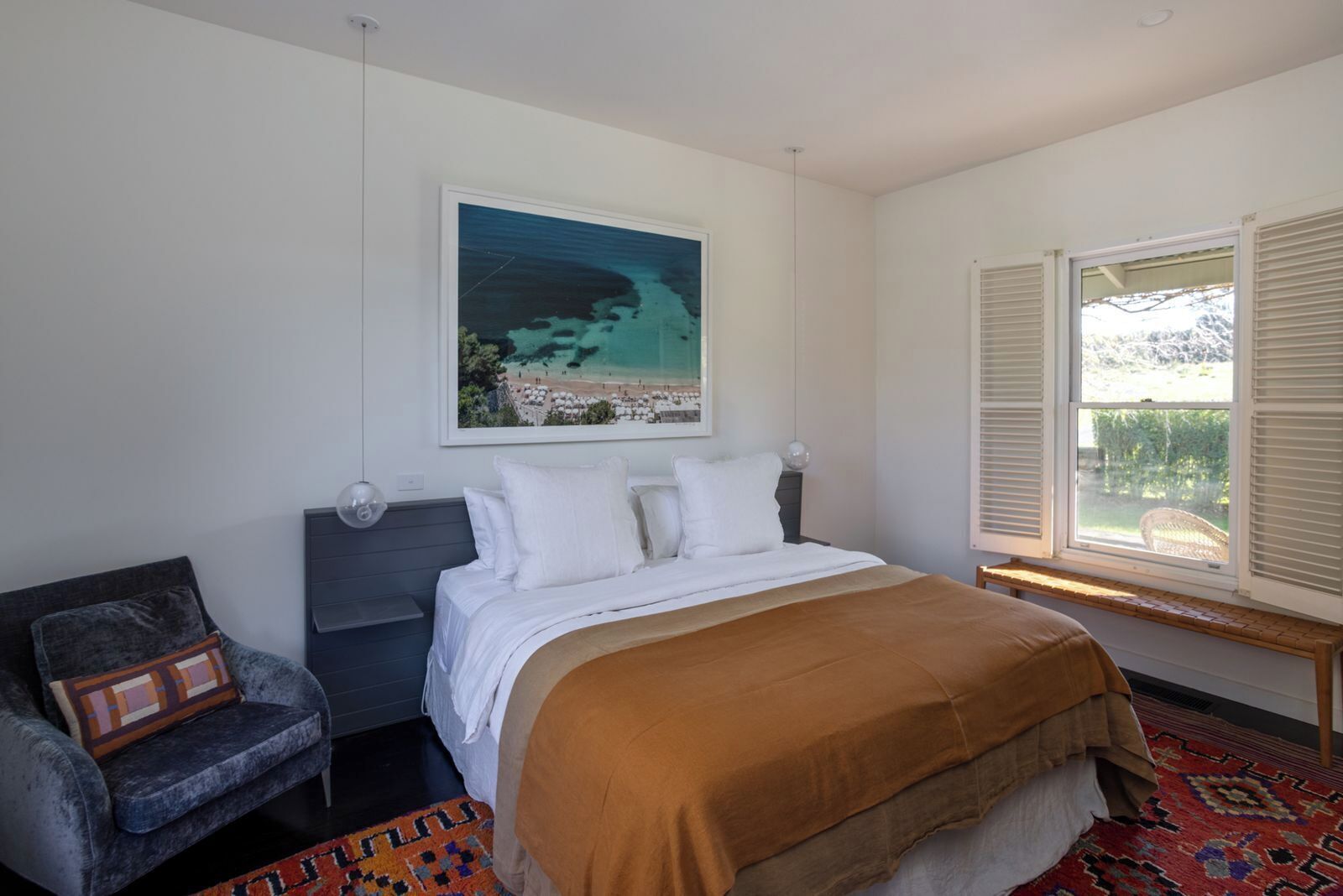 A bedroom with a bed, a chair, a window with blinds, and a painting of a beach scene.