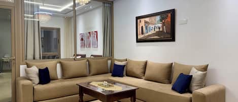 A living room with a sectional sofa, a wooden coffee table, a chandelier, a framed picture, and a large window with curtains.