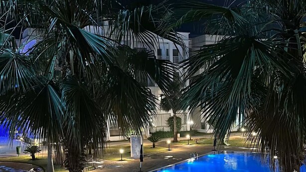 Un área de piscina iluminada de noche, con palmeras y un edificio al fondo.