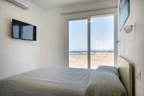 A bedroom with a bed, a TV, a sliding glass door to the beach, and a small wall-mounted shelf. - Green Oasi 1 Marzamemi  (Marzamemi)