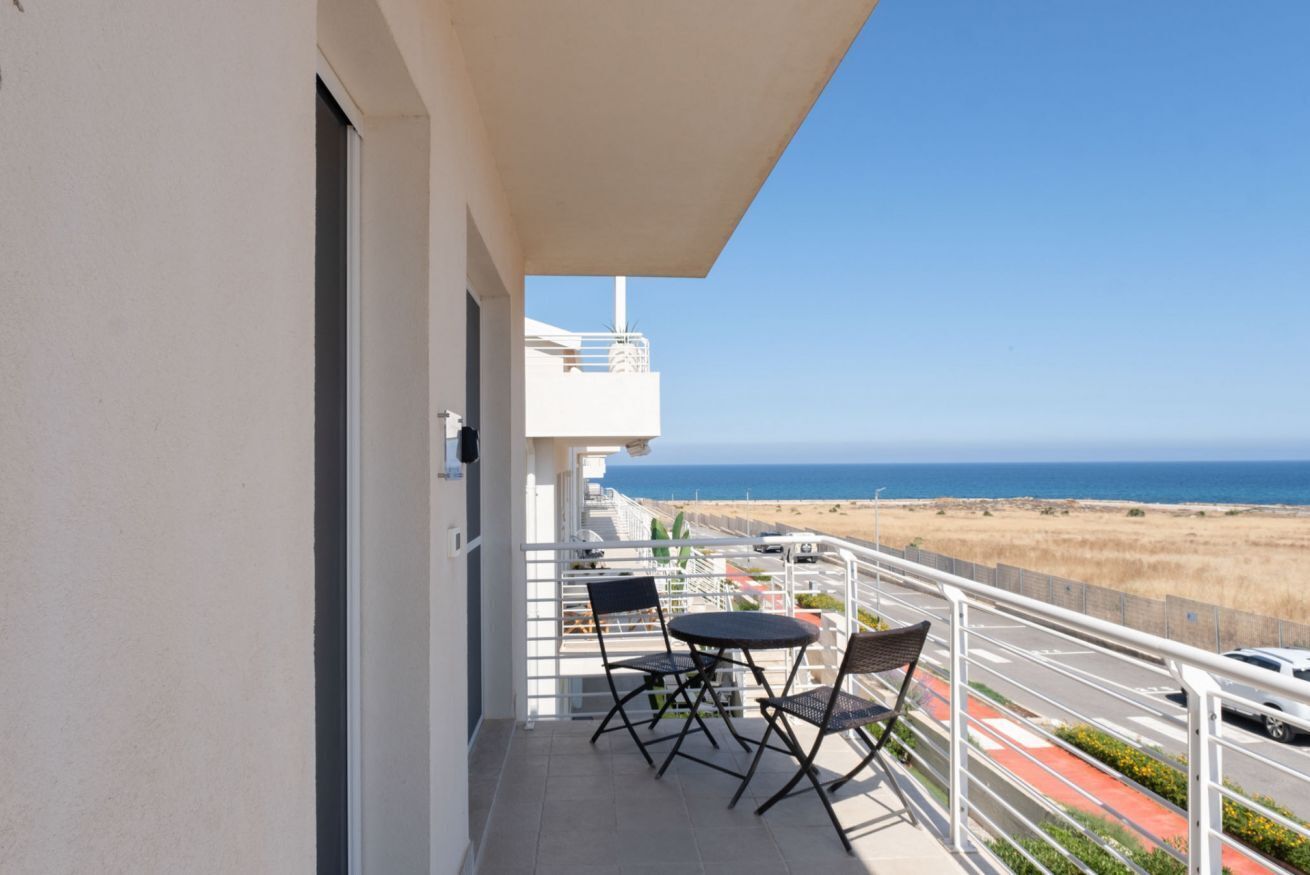 A balcony with a table and chairs, a view of the beach, and a clear sky.
