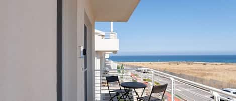 A balcony with a table and chairs, a view of the beach, and a clear sky.