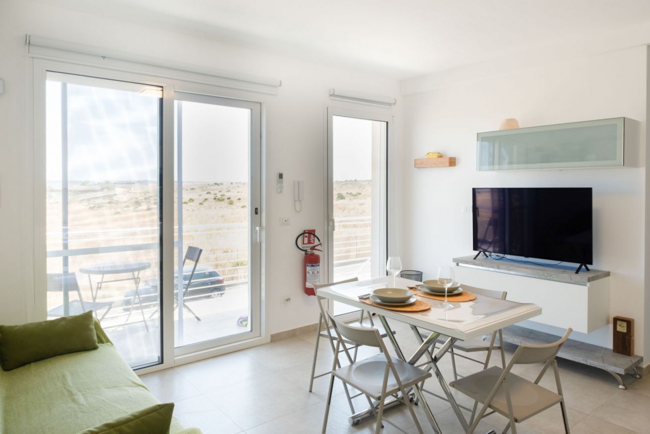 A modern room with a dining table, chairs, a sofa, a TV, and a sliding glass door leading to an outdoor area.