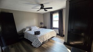 Ein Schlafzimmer mit einem Bett, einem Nachttisch, einem Deckenventilator, einem Fenster mit Vorhängen und einer dunklen Tür.