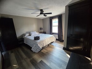 A bedroom with a bed, a nightstand, a ceiling fan, a window with curtains, and a dark door. - A Hop ,Skip, and Jump to the Beach (Lorain)