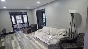 Ein Wohnzimmer mit einer Couch, zwei Sesseln, einem Couchtisch und einer Stehlampe. Im Hintergrund ist eine Essecke mit Tisch und Stühlen zu sehen.