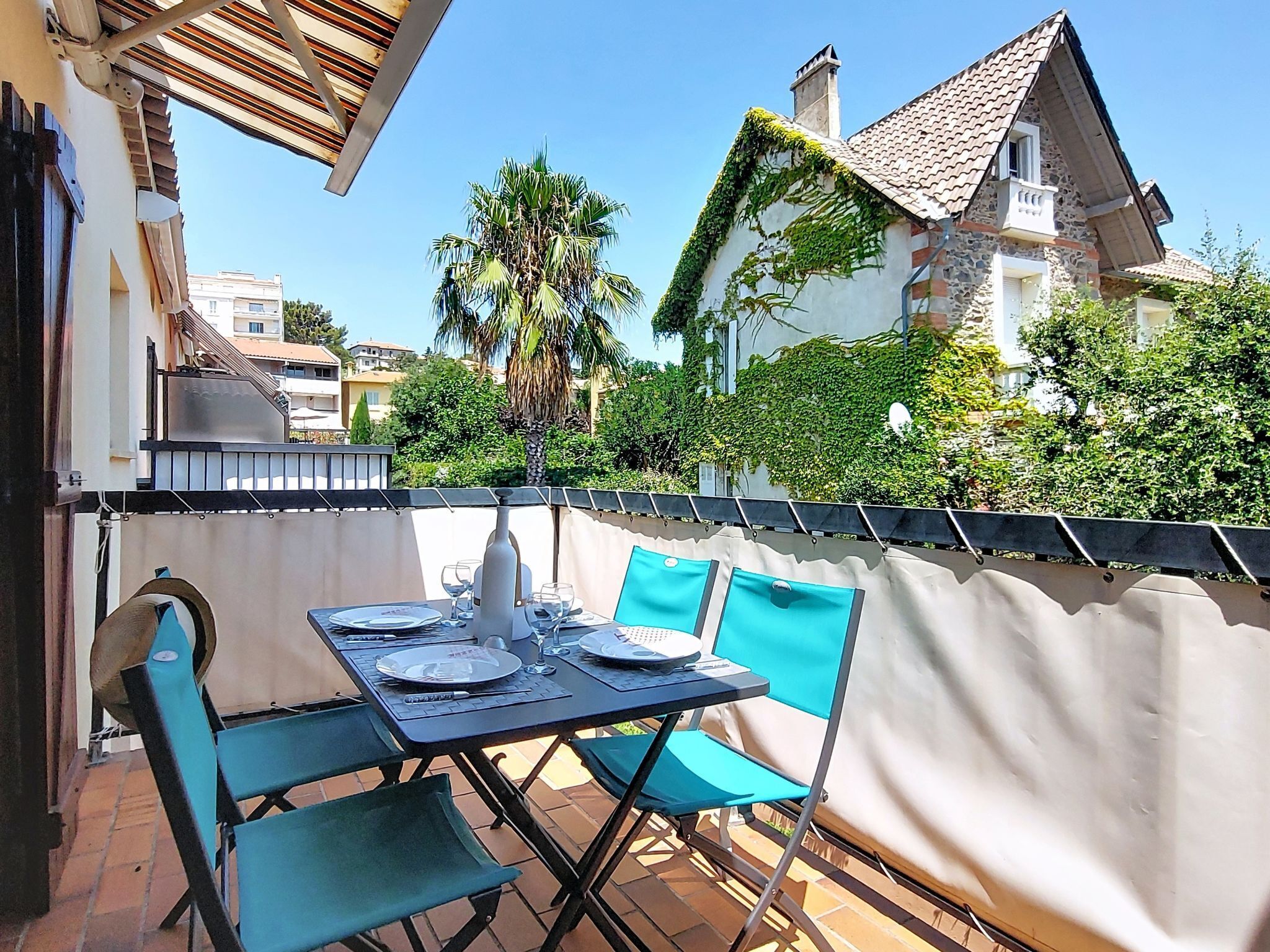 An outdoor patio with a table set for two, surrounded by a white fence, with buildings and greenery in the background.