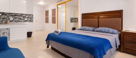 Un dormitorio con cama, mesitas de noche, cabecera de madera, espejo, baño y kitchenette.