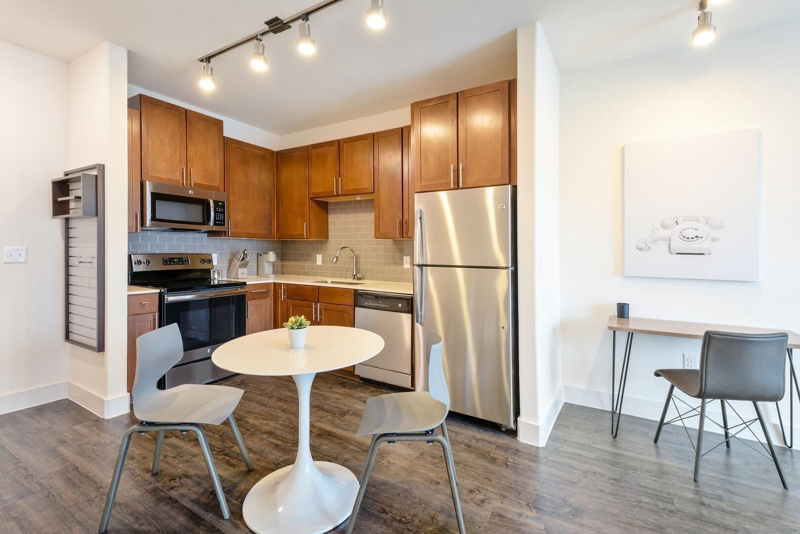 A modern kitchen with wooden cabinets, stainless steel appliances, a dining table with chairs, and a small office area with a desk and chair.