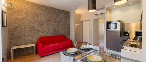A compact living space with a dining area, a red sofa, a kitchen with modern appliances, and a neutral-toned wall.