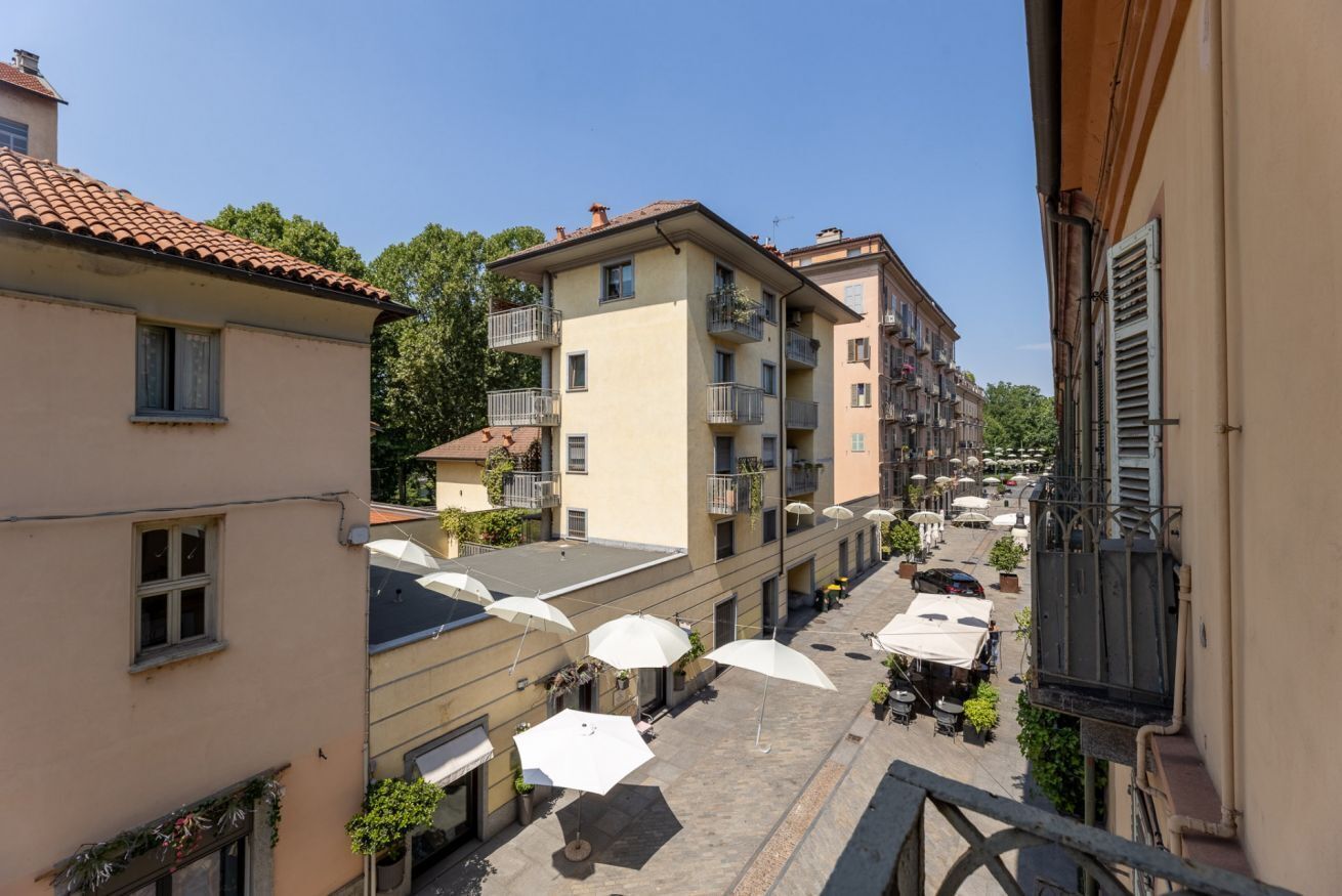 Une rue étroite, bordée de bâtiments des deux côtés, avec des tables et des chaises installées à l’extérieur sous des parasols, et des balcons dotés de balustrades.